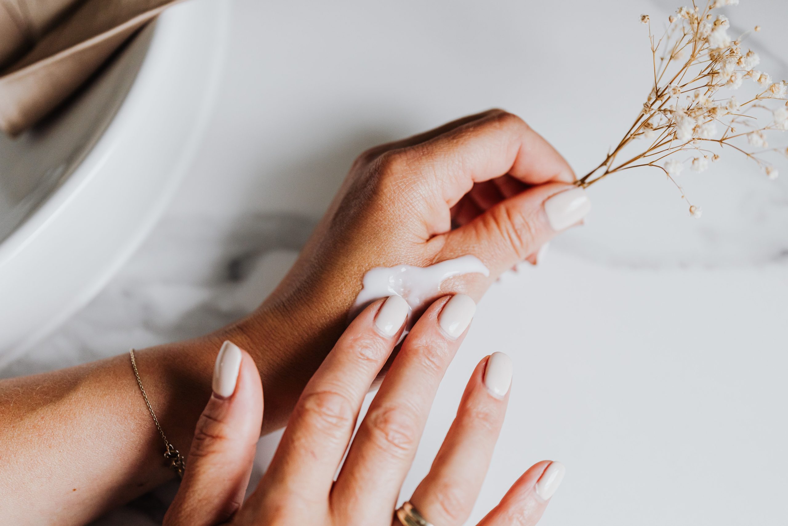 Close-up of manicured hands applying cream with a delicate floral accent on a marble surface.